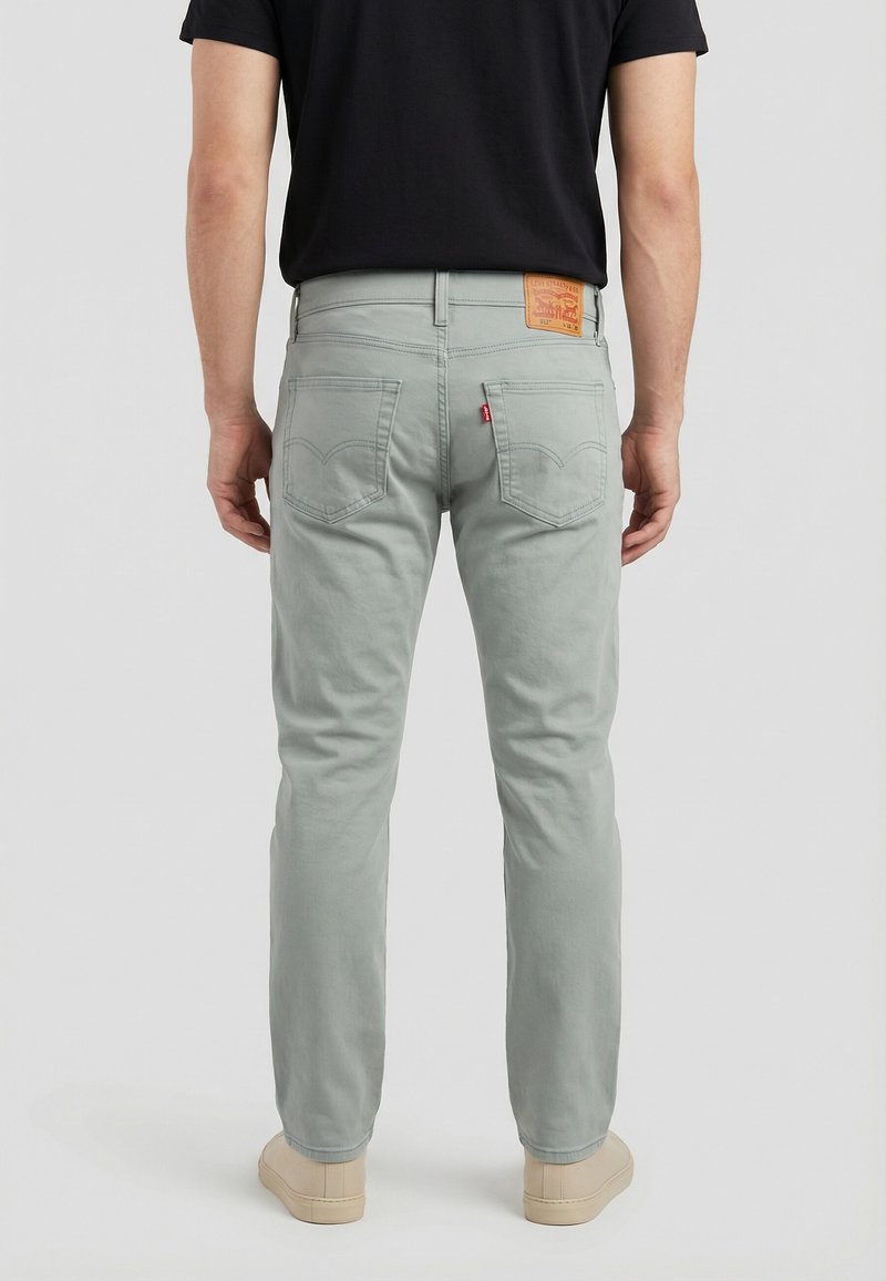 Hombre con vaqueros gris claro con bolsillos traseros y una camiseta negra, de pie frente a un fondo liso y claro.