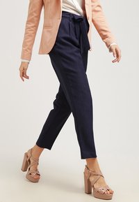 Námořnické kalhoty na míru s páskem v pase, doplněné světle růžovými sandály na vyšším podpatku. Oblečení se vyznačuje hladkým materiálem a slim fit střihem.