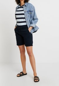 Kurtka dżinsowa, niebiesko-biała koszula w paski, ciemne szorty i czarne klapki z srebrnymi akcentami. Casualowy strój odpowiedni na ciepłą pogodę.