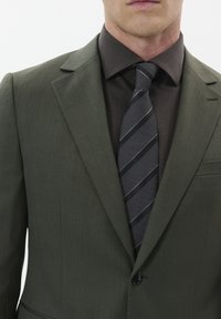 Alyvuogių žalios spalvos švarkas su ryškiu apykaklė, derinamas su tekstūruota tamsiai ruda marškinėliais ir juoda kaklaraiščiu su pilkomis juostelėmis.