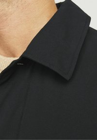 Černý kabát se strukturovaným límcem, který má hladkou texturu a čisté linie. Tkanina se jeví jako lehká a voděodolná.