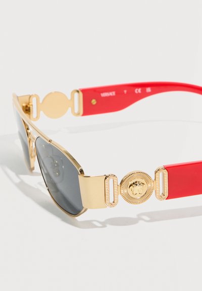Γυαλιά ηλίου με χρυσή κορνίζα, σκούρους φακούς και κόκκινα μπράτσα που φέρουν χρυσό έμβλημα Medusa στις μεντεσέδες.