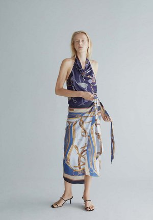 Mujer de pie en el interior con un top halter azul marino y una falda cruzada con estampado azul, blanco y dorado, y sandalias negras de tacón.
