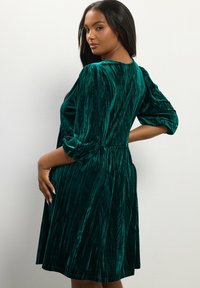 Smaragdgrünes Samtkleid mit dreiviertel Ärmeln, A-Linienform und gerafftem Taillenbereich. Das Kleid zeichnet sich durch eine weiche Textur und eine fließende Silhouette aus.