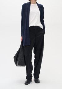 Cárdigan largo azul marino sobre una camisa blanca, combinado con pantalones negros de cintura alta y mocasines negros. Presenta un bolso tote negro.