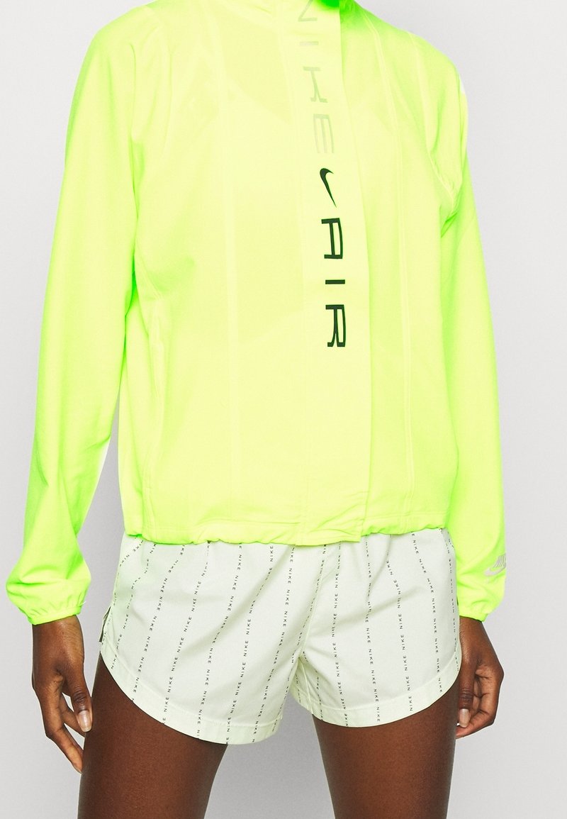 Giacca a vento gialla neon con dettagli logo e zip frontale, abbinata a pantaloni corti verdi chiari con branding ripetuto e un fit rilassato.