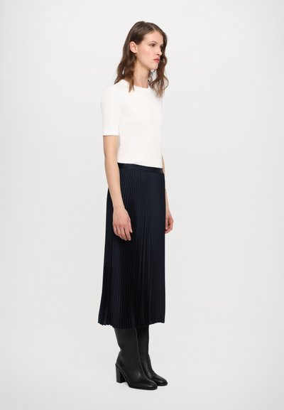 Top bianco a maniche corte, gonna a pieghe blu navy, stivali neri al ginocchio con tacco. Tessuto liscio con pieghe verticali, vita aderente, silhouette elegante.