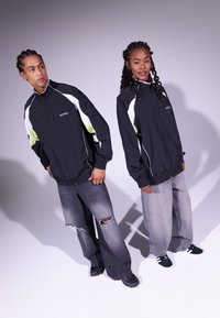 Casacos pretos com detalhes em branco e verde neon, jeans de corte relaxado e sapatilhas pretas. Estilo casual de streetwear.