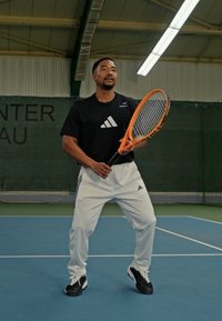 Camiseta deportiva negra con el logo blanco de adidas, pantalones blancos con rayas negras, sosteniendo una raqueta de tenis naranja en una cancha azul. Entorno interior.