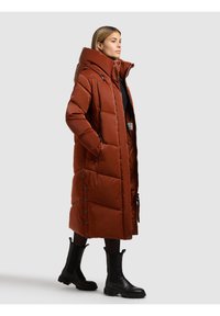 Mujer con abrigo acolchado largo de color óxido con cuello alto y botas negras, de pie de perfil sobre un fondo liso.