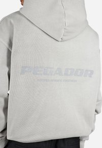 Sudadera con capucha gris de tela texturizada, que presenta un gran logo "PEGADOR" en azul claro en la parte posterior y un diseño minimalista.