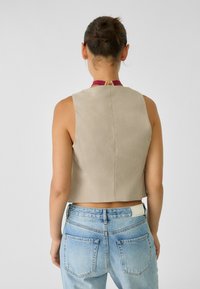 Beżowy prążkowany crop top z wysokim dekoltem, z bordowym kołnierzykiem i złotym zapięciem z tyłu, w połączeniu z niebieskimi dżinsami z wysokim stanem.