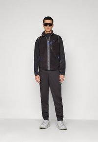 Man wearing black fleece vest over long-sleeve top, black track pants, white sneakers, and large black sunglasses standing against plain background.