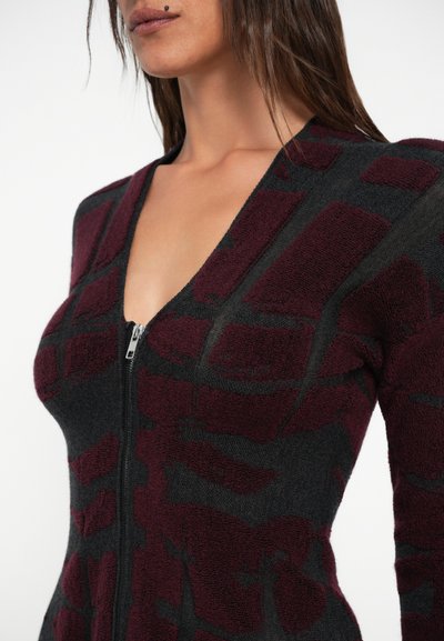Crna i burgundska zip-up dukserica s teksturiranim uzorkom, koja ima duboki V-izrez i geometrijske oblike. Materijal izgleda mekan i pahuljast.