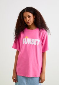 Růžové bavlněné tričko s nápisem "SUNSET" velkými bílými písmeny a symbolem žlutého slunce, volný střih s krátkými rukávy.