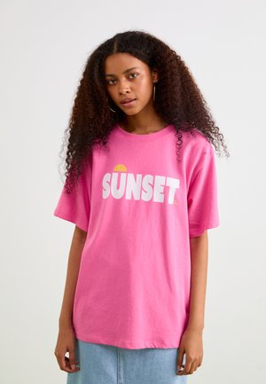 Vaaleanpunainen puuvillapaita, jossa on suuri valkoinen teksti "SUNSET" ja keltainen auringonsymboli. Rentoa mitoitusta, jossa on lyhyet hihat.