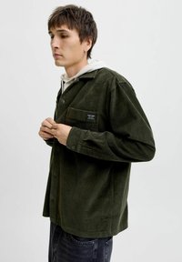 Donkergroene corduroyshirt met een knoopsluiting, borstzak en gestructureerde stof. Gedragen over een witte hoodie.