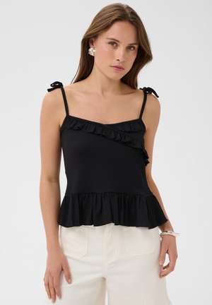Femme portant un haut noir sans manches avec des volants et un pantalon blanc, debout devant un fond clair uni.
