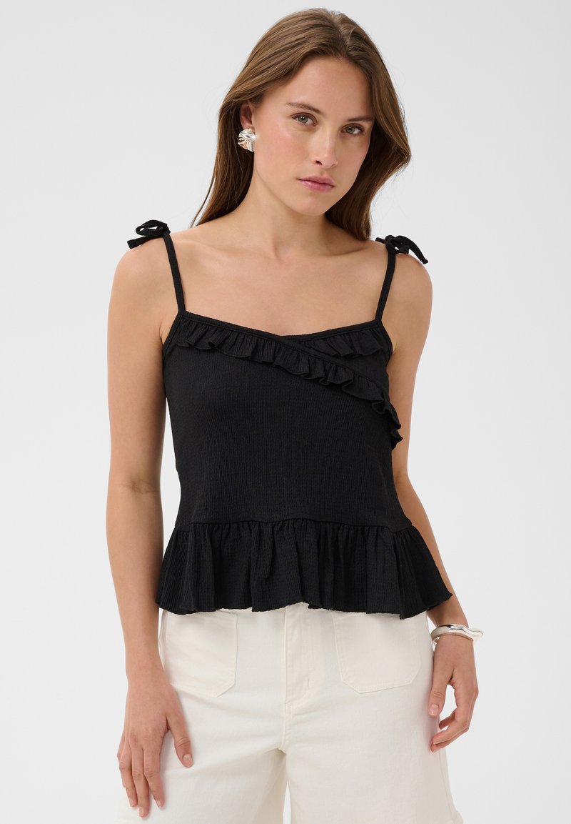 Femme portant un haut noir sans manches avec des volants et un pantalon blanc, debout devant un fond clair uni.