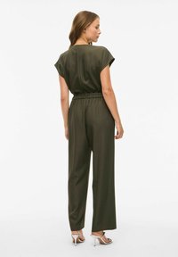 Mujer de pie, vistiendo un mono corto de manga corta en verde oliva con cinturón y pantalones de pierna ancha, con sandalias de tacón alto plateadas, de espaldas.