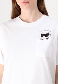 Bílý bavlněný tričko s kulatým výstřihem a krátkými rukávy, s grafikou postavy se slunečními brýlemi na náprsním kapsičce.