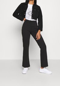 Chaqueta de mezclilla negra recortada con botones plateados, camiseta gráfica blanca, pantalones ajustados negros y zapatillas blancas. Diseño simple y aerodinámico.