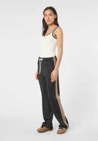 Mujer de pie mirando ligeramente de lado, vestida con camiseta sin mangas blanca, pantalones negros con franjas laterales beige y zapatos con estampado de leopardo sobre un fondo liso.
