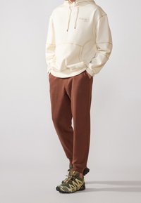 Cremefärgad hoodie med framficka och brodyr, i kombination med bruna joggerbyxor och gröna, svarta och tanfärgade sportskor.