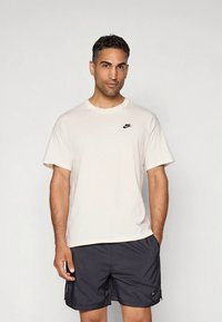 Hellbeige Nike-T-Shirt mit kurzen Ärmeln, rundem Ausschnitt und kleinem schwarzem Logo auf der linken Seite, kombiniert mit schwarzen Shorts.
