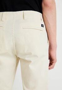 Pantalonii bej din bumbac au o croială dreaptă, două buzunare la spate și un eticheta vizibilă a mărcii pe talie. Textură netedă.
