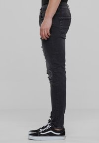 Jeans ajustados negros rasgados con una textura deslavada, que presentan detalles deshilachados en las rodillas, desgastados en el dobladillo, combinados con zapatillas negras de skate.