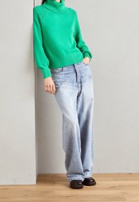 Pull vert tricoté à col montant, manches texturées, associé à un jean large bleu clair et des chaussures noires, appuyé contre un mur.