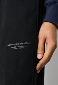 Ręka obok czarnej tkaniny z białym nadrukiem tekstu "Abercrombie and Fitch Założona w Nowym Jorku Wysokiej Jakości Towary".