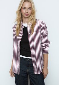 Chemise à rayures boutonnée bordeaux et blanche, avec une poche poitrine et des manches retroussées, portée sur un haut noir et un jean taille haute foncé.