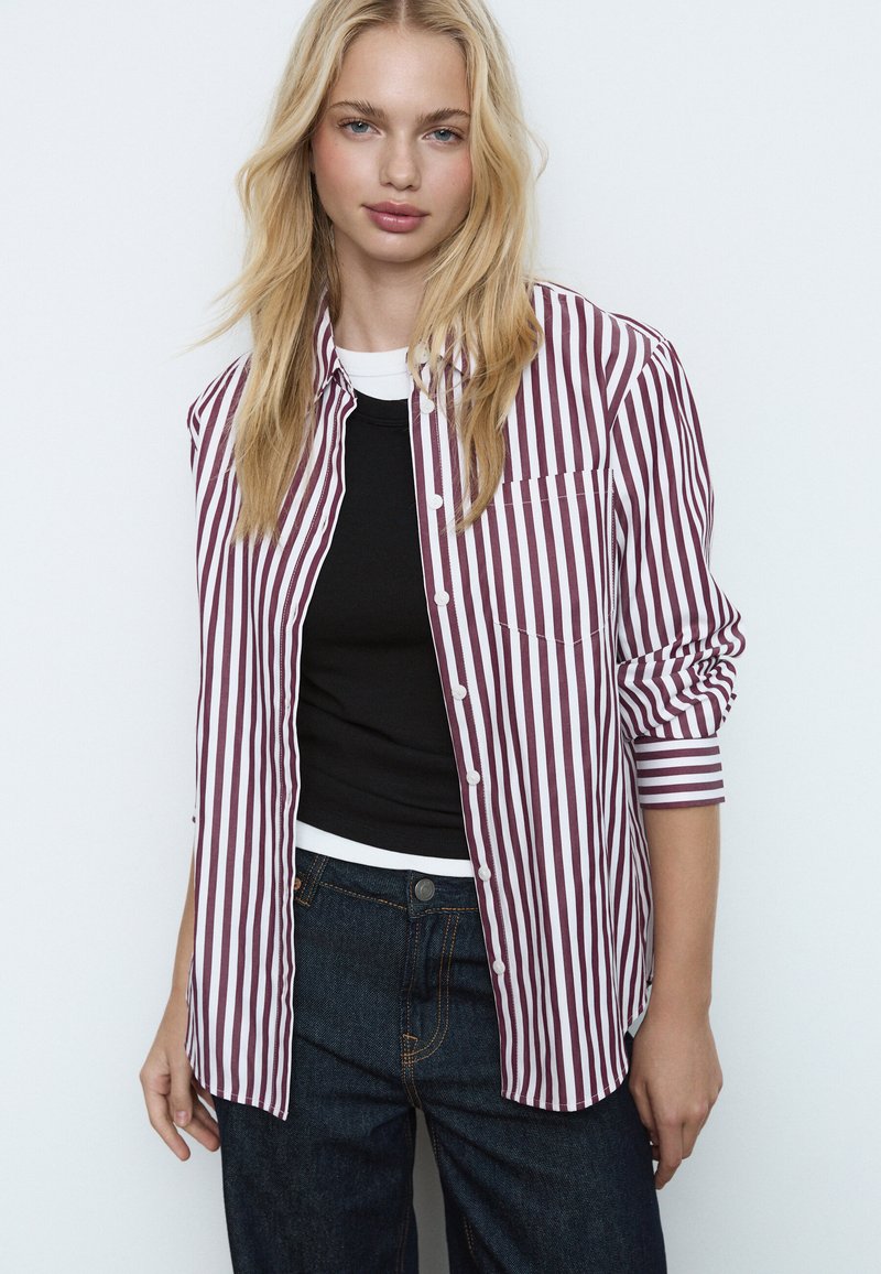 Chemise à rayures boutonnée bordeaux et blanche, avec une poche poitrine et des manches retroussées, portée sur un haut noir et un jean taille haute foncé.