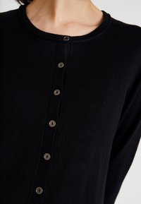 Sort cardigan lavet af bl�ødt materiale med rund halsudskæring, ribbet detalje og seks metalliske knapper foran.