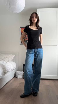 Junge Frau steht im Schlafzimmer, trägt ein schwarzes eng anliegendes Oberteil, weite blaue Jeans und schwarze Schuhe, die Hände hinter dem Rücken verschränkt.