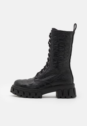 Botte de combat noire à lacets avec motif en fausse peau de crocodile texturée en relief et semelle épaisse à tranchant dentelé, vue de côté sur fond blanc.
