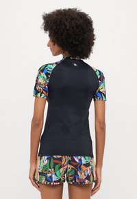 T-shirt z krótkim rękawem, w czarnym kolorze z kolorowymi rękawami w kwiatowy wzór. Może być zestawiony z dopasowanymi szortami pływackimi w kwiaty. Gładka struktura materiału.