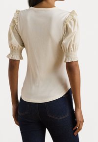 Kvinna som bär en krämfärgad ribbad topp med puffiga, volangärmshorts och mörkblå jeans, sedd från baksidan.