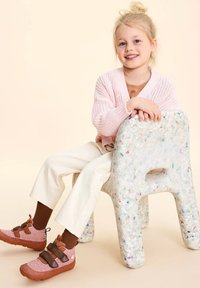 Scarpe per bambini: tomaia in tessuto rosa con cinturini marroni, poggiate su una sedia in plastica riciclata. Pantaloni bianchi e un cardigan lavorato a maglia rosa chiaro.