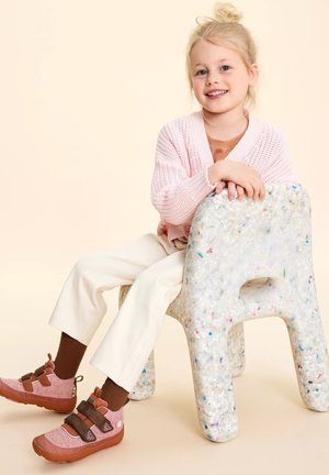 Scarpe per bambini: tomaia in tessuto rosa con cinturini marroni, poggiate su una sedia in plastica riciclata. Pantaloni bianchi e un cardigan lavorato a maglia rosa chiaro.