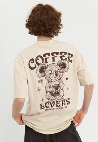 Beige oversized t-shirt med et brunt grafik af en koala, der holder en kaffekop, med "COFFEE LOVERS" ovenfor og "ESPRESSO YOURSELF" nedenfor.
