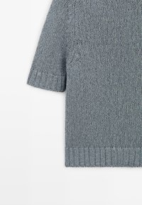 Lysegrå strikket trøje med korte ærmer, der viser ribstrikket kant og manchetter, tekstureret strikkemønster samt et close-up af skulder og ærme.