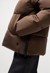 Brun pufferjakke med quiltet design, der har sorte lynlåse og lommeaccenter. Teksturen er glat med en let blank finish.