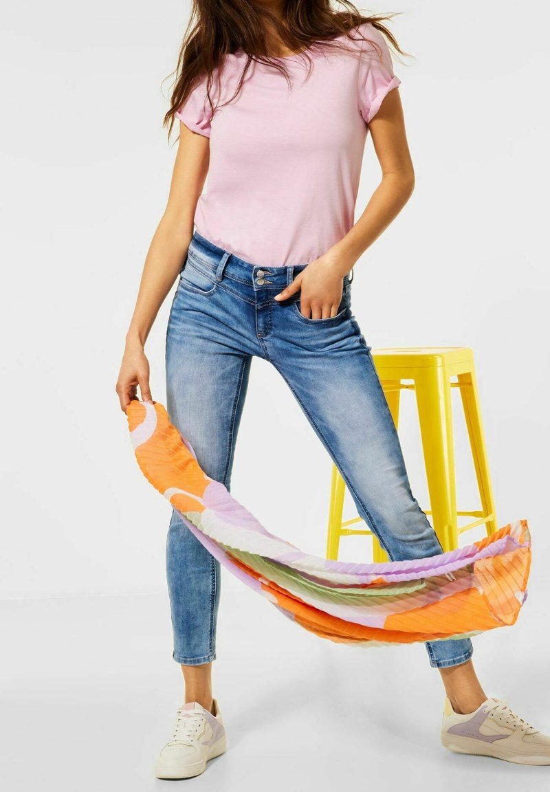 Růžové přiléhavé tričko, světle modré skinny džíny, bílé tenisky, držící barevně vzorovaný šátek v oranžové, růžové a zelené. Žluté stoličky v pozadí.