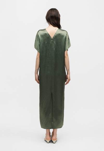 Γυναίκα που φορά ένα φαρδύ, λαμπερό μεσαίο φόρεμα σε ελιά πράσινο χρώμα με κοντά μανίκια, λαιμόκοψη V στην πλάτη και σκίσιμο στο κέντρο της πλάτης, στέκεται σε απλό λευκό φόντο.