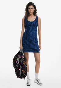 Rochie din denim albastru cu un design ajustat, având un material texturat, cusături în contrast și un decolteu rotund. Modelul ține un rucsac cu motive florale.