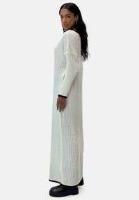 Langes, weißes Strickkleid mit vertikalen Rippen und langen Ärmeln. Schwarz akzentuierte Bündchen und Saum, kombiniert mit klobigen schwarzen Stiefeln.
