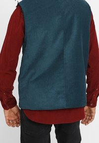 Mann, der eine türkisfarbene ärmellose Jacke über einem langärmeligen roten Hemd und schwarzen Hosen trägt, von hinten gesehen.